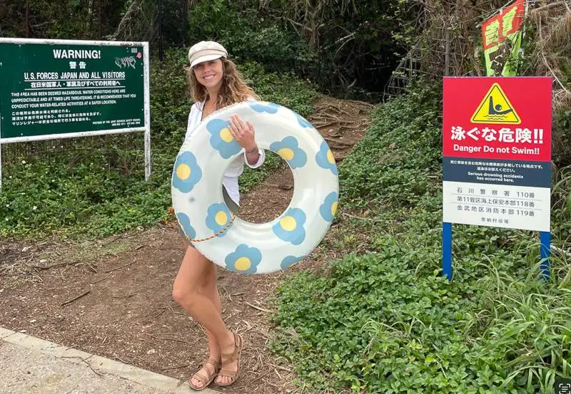 Girl holding an innertube in front of signs that say 'no swimming' and 'danger, do not enter water' in Okinawa, Japan