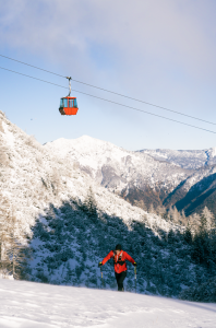 A man in a red jacket hiking up a snow covered mountain, with a red cable car overhead